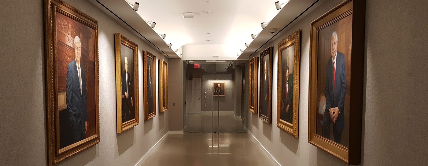 A well-lit, narrow hallway lined on both sides with multiple large, framed, gold-framed oil portraits of professional men, leading toward a glass door at the end.