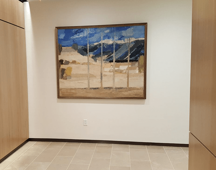 A large, framed landscape painting in a semi-abstract style, featuring blue skies, distant mountains, and light-colored trees against a tan and brown foreground, hanging on a white wall.